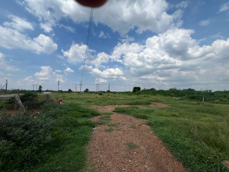 A view of a vacant residential plot featuring natural grass cover, wire fencing, and overhead power lines under a cloudy sky.