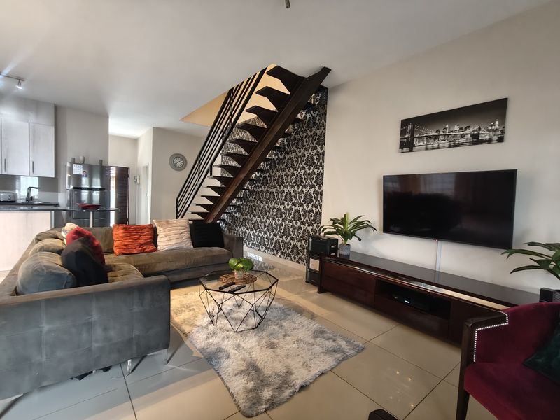 An open-plan living area featuring a modern open-tread staircase, tiled flooring, and a decorative feature wall, seamlessly connected to the kitchen space.