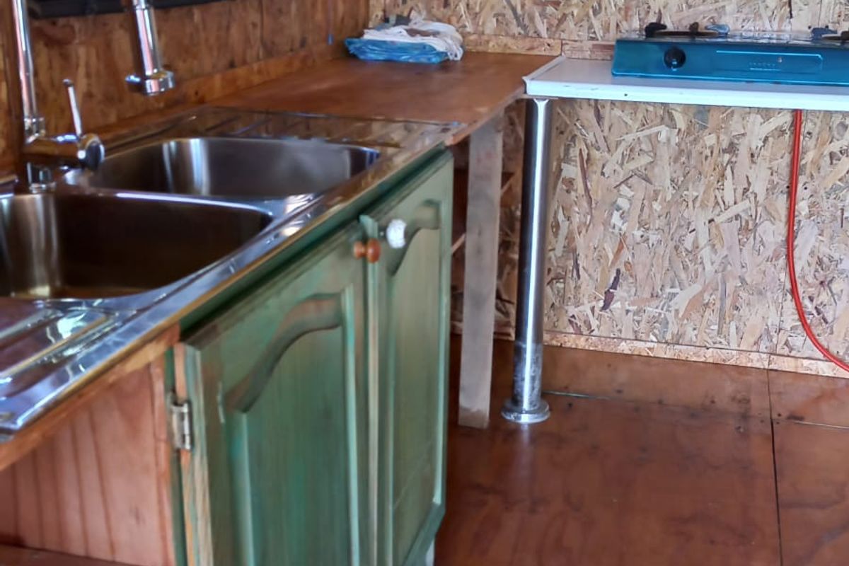 A rustic kitchen area featuring a double stainless steel sink, wooden cabinetry, and OSB wall paneling with a portable gas cooktop.