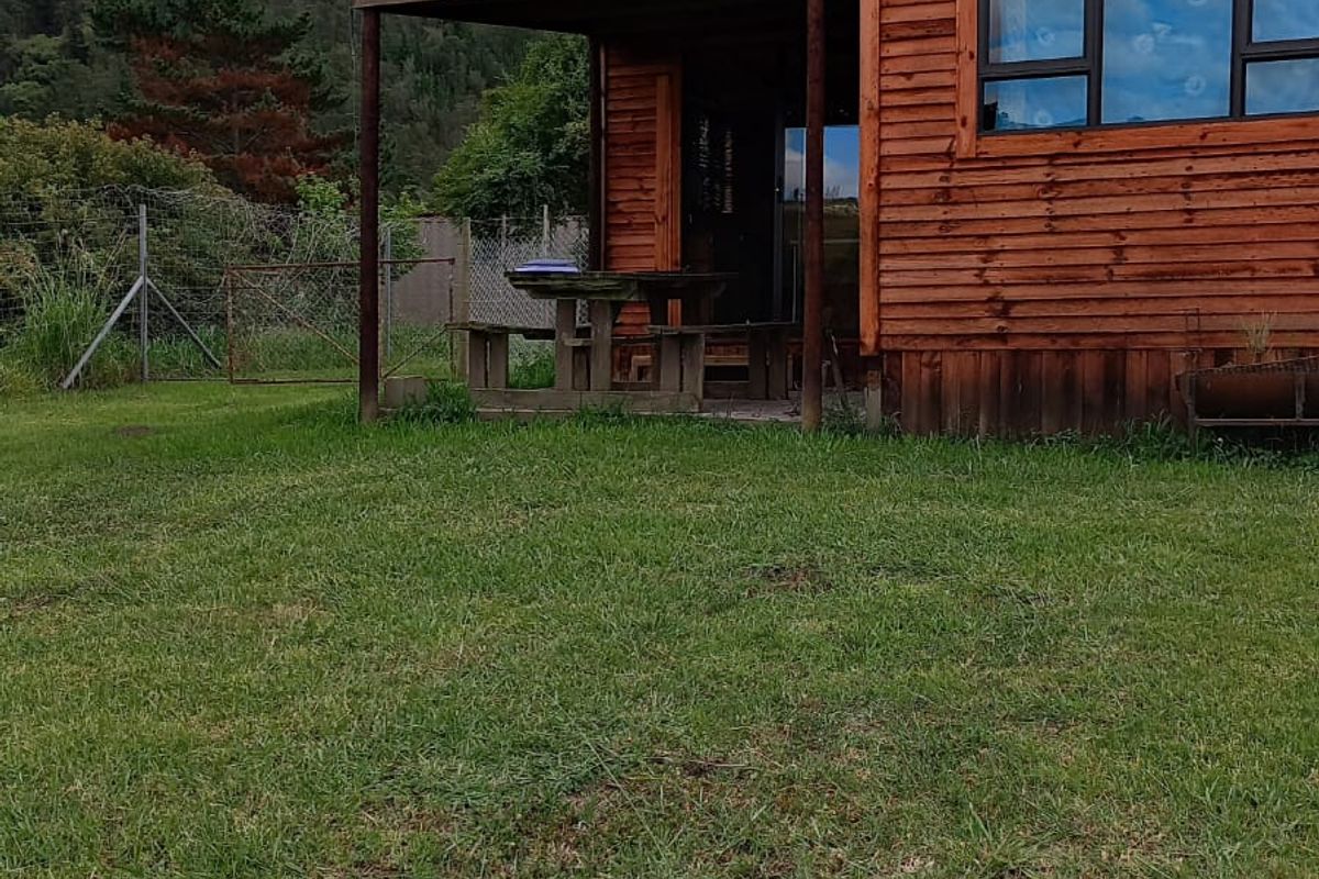A timber-clad cabin featuring a covered patio area, set against a scenic mountain backdrop with expansive lawn space.