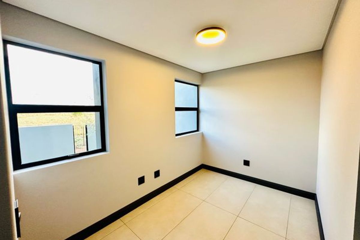 A bright, modern, and empty bedroom featuring neutral tiled flooring, dark window frames, and a contemporary flush-mount ceiling light.