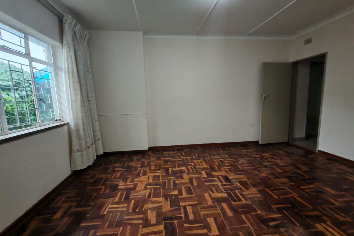 An empty bedroom featuring classic parquet flooring, large windows with security bars, and neutral wall finishes, offering a blank canvas for personalization.