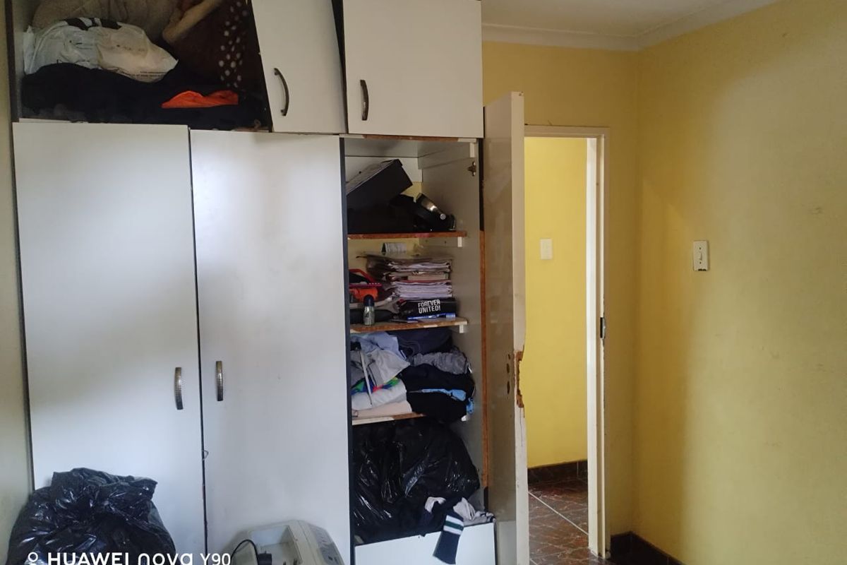A bedroom featuring built-in cupboards with visible wear and tear, leading to an adjacent hallway with tiled flooring.