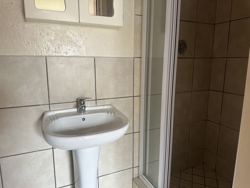 A functional bathroom featuring a pedestal washbasin, a shower enclosure, and wall-mounted storage, finished with neutral ceramic tiling.