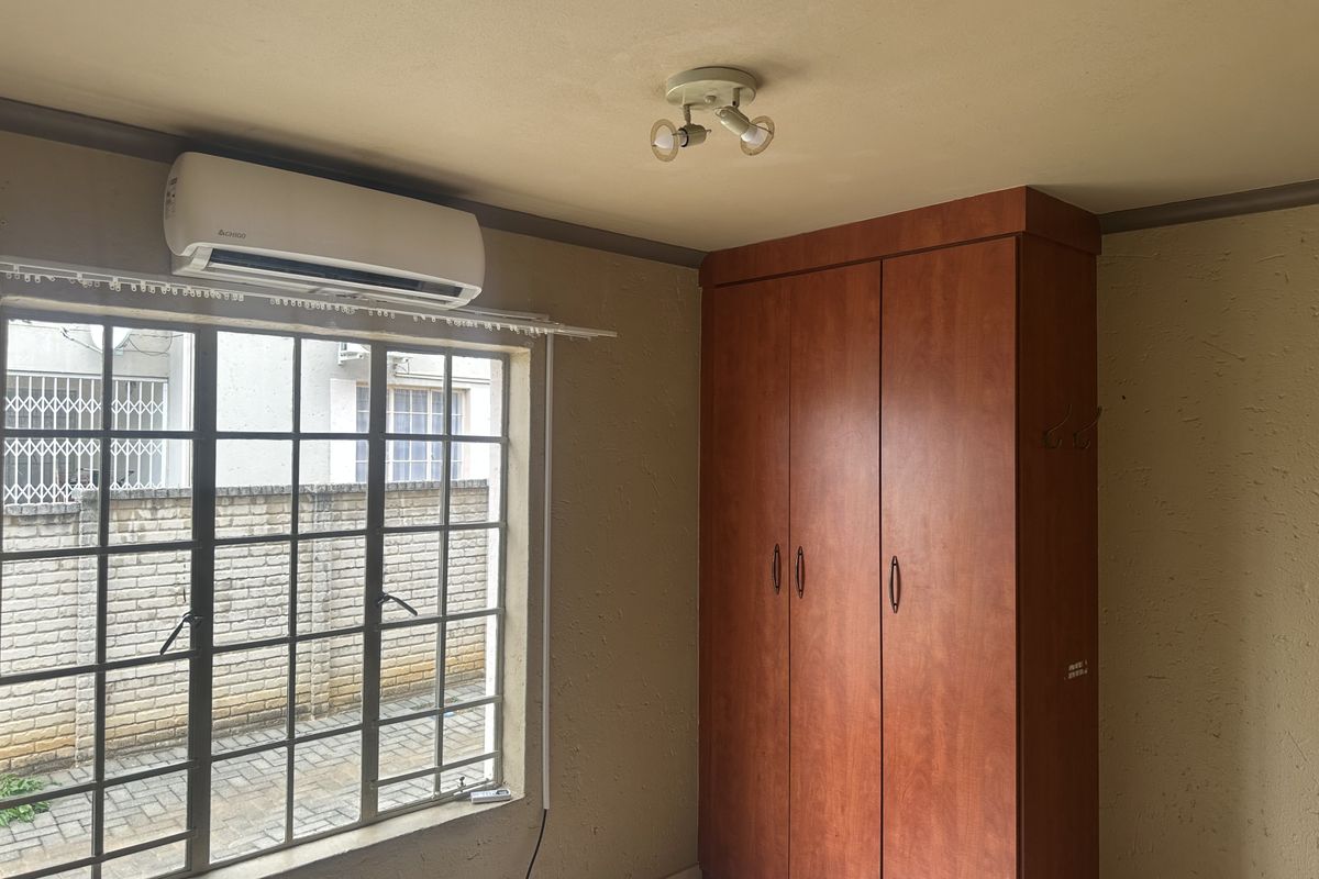A bedroom featuring a built-in wardrobe, air conditioning unit, and security bars on the window, with tiled flooring throughout.