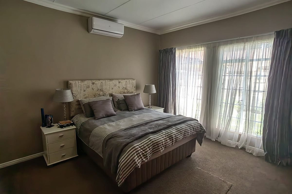 A well-maintained bedroom featuring neutral wall tones, carpeted flooring, a wall-mounted air conditioning unit, and sliding glass doors providing natural light.