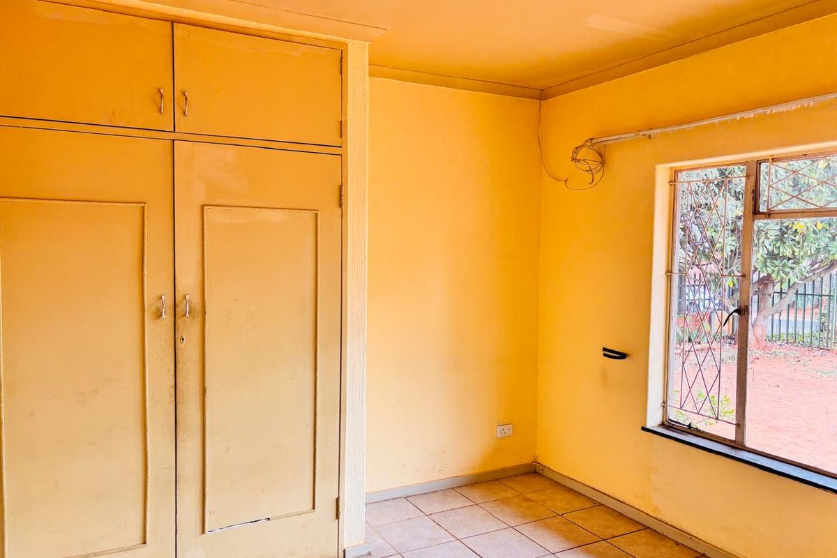 A bedroom featuring built-in cupboards, tiled flooring, and a window with security bars, offering a blank canvas for renovation.