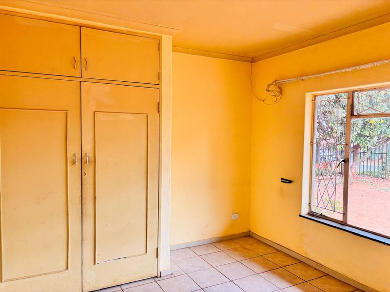 A bedroom featuring built-in cupboards, tiled flooring, and a window with security bars, offering a blank canvas for renovation.