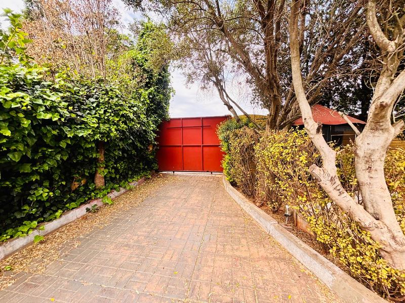 A paved driveway leading to a large red metal gate, framed by mature trees and dense garden hedging.