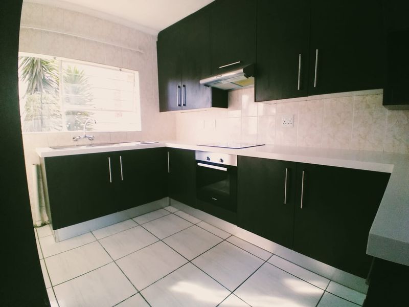 A clean, modern kitchen featuring dark cabinetry, white countertops, built-in oven and hob, and tiled flooring with ample natural light.