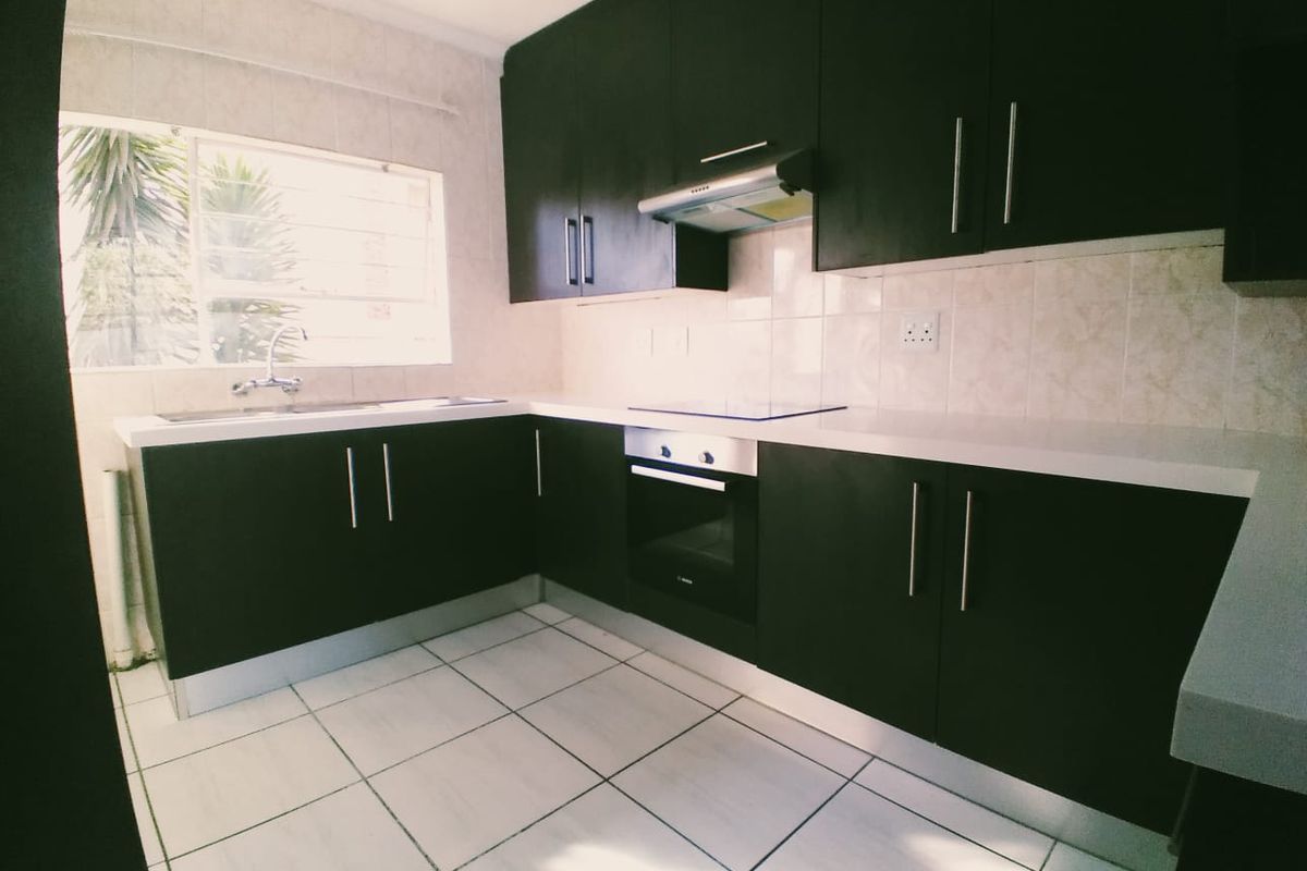 A clean, modern kitchen featuring dark cabinetry, white countertops, built-in oven and hob, and tiled flooring with ample natural light.