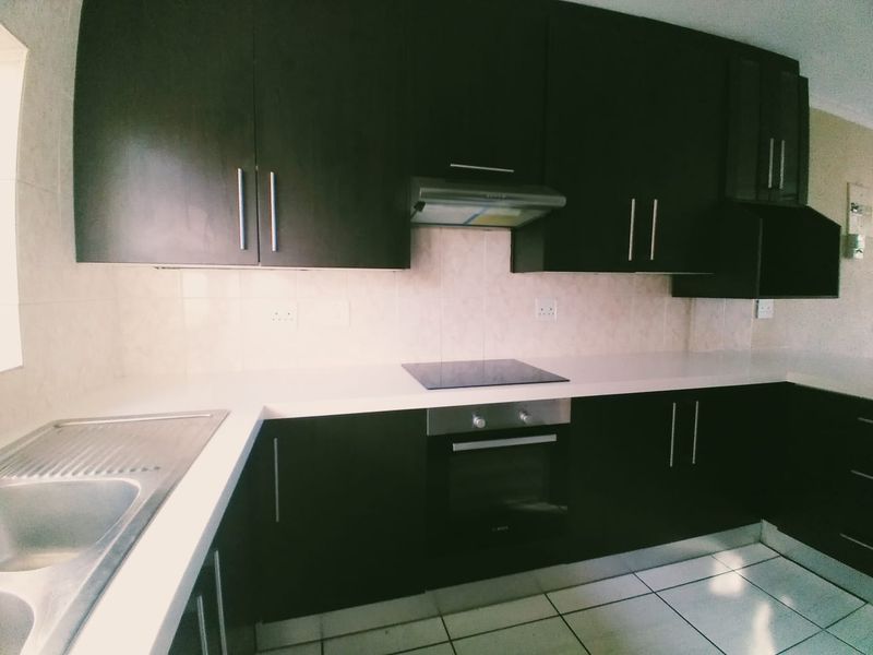 Modern kitchen featuring dark cabinetry, white stone countertops, built-in oven, electric hob, and stainless steel sink with tiled backsplash and flooring.