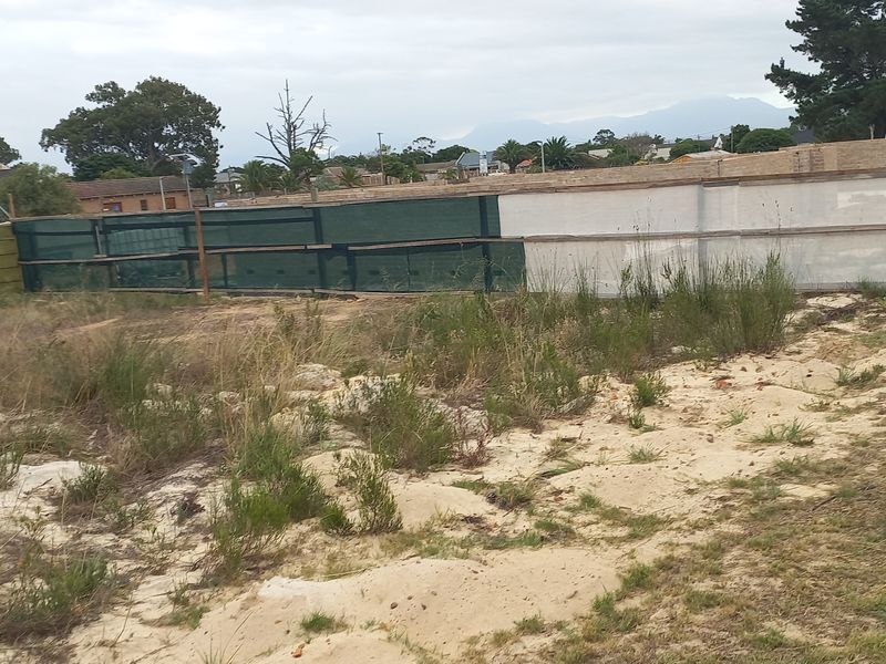 A vacant residential plot featuring sandy terrain, overgrown vegetation, and temporary boundary fencing, situated in a developed neighborhood.