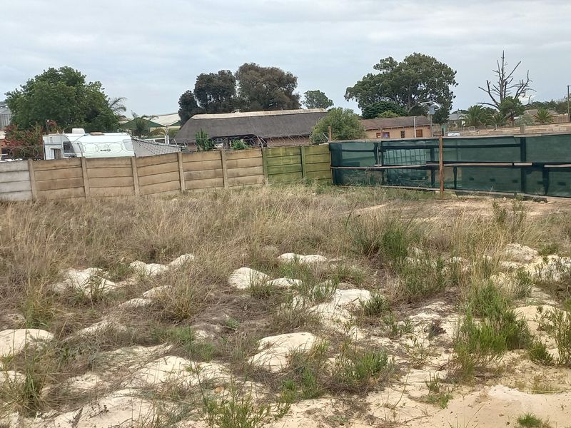 A vacant residential plot featuring sandy terrain, overgrown grass, and existing boundary walling with shade cloth screening.