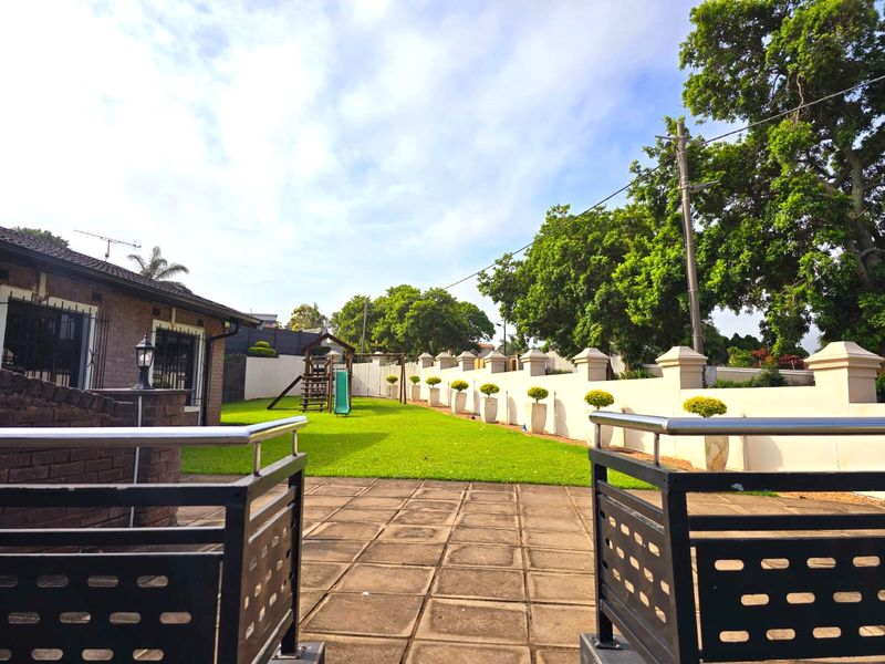 A well-maintained rear garden featuring a paved patio area, metal balustrades, and a manicured lawn enclosed by a perimeter wall.