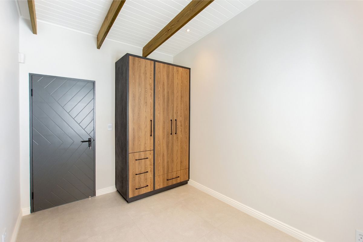 A bright, modern bedroom featuring high ceilings with exposed timber beams, tiled flooring, and a contemporary built-in wardrobe with wood-grain finish.