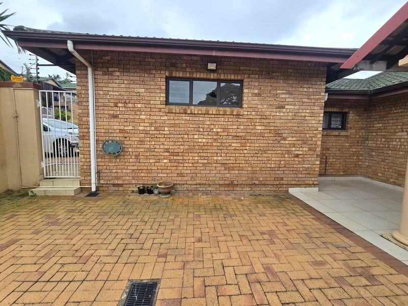 A rear exterior view featuring a face brick facade, paved driveway with drainage, and a small tiled patio area.