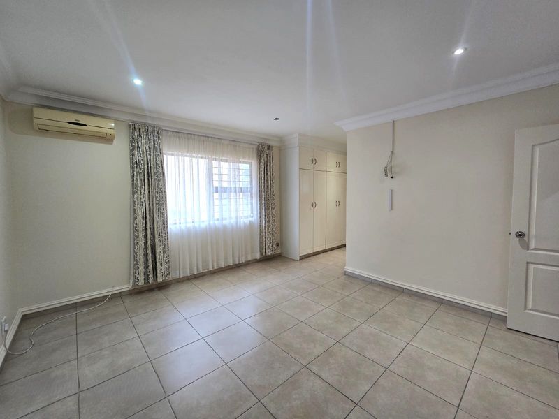 A spacious, neutral bedroom featuring tiled flooring, built-in cupboards, recessed lighting, and an installed air conditioning unit.