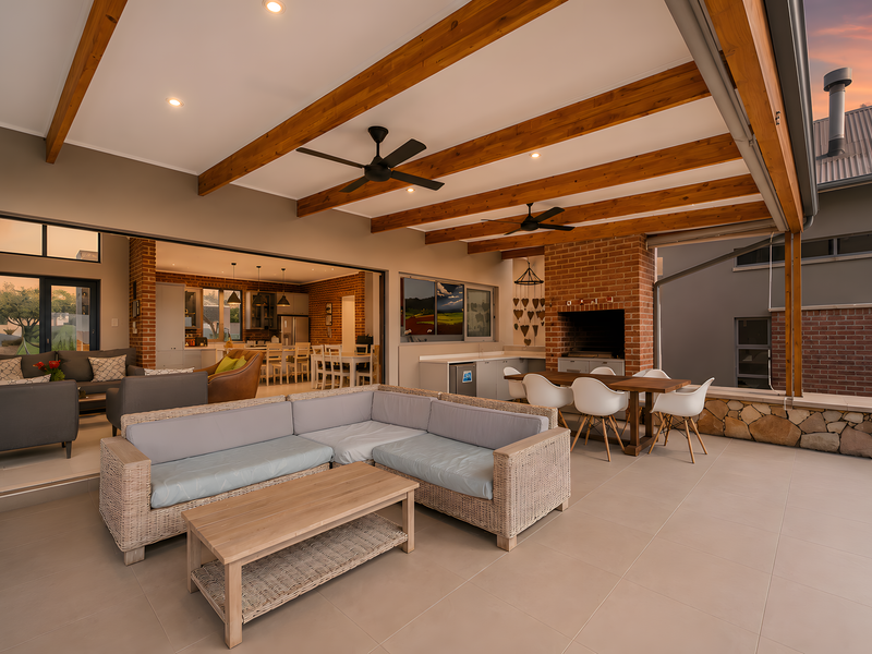 Spacious covered patio featuring exposed timber beams, a built-in brick fireplace, and seamless flow to the open-plan interior living and kitchen area.