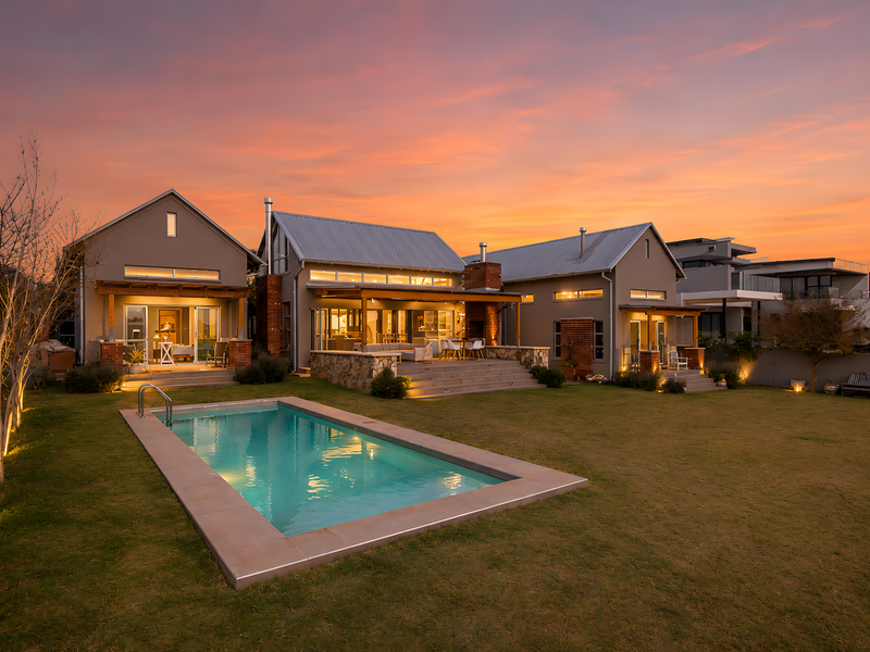Stunning rear exterior view of a modern home featuring a swimming pool, stone-clad patio, outdoor fireplace, and beautifully landscaped garden at sunset.