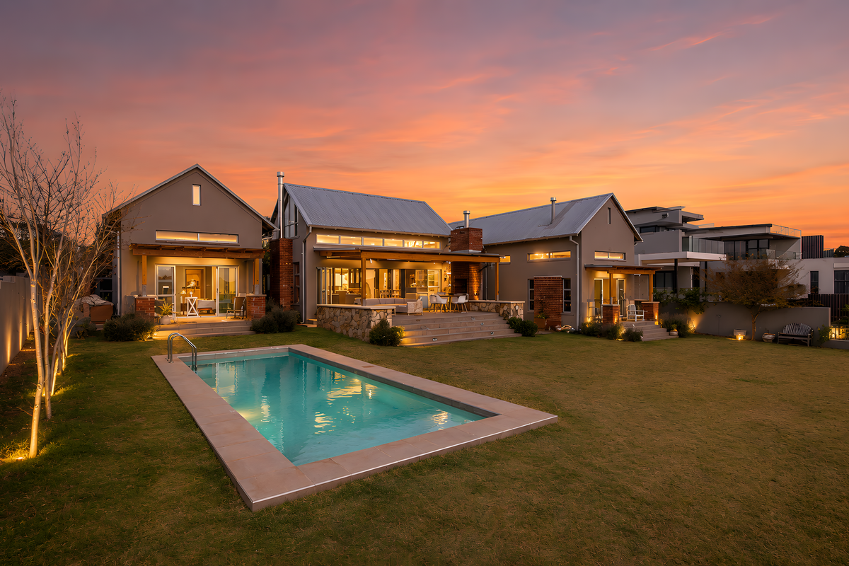Stunning rear exterior view of a modern home featuring a swimming pool, stone-clad patio, outdoor fireplace, and beautifully landscaped garden at sunset.