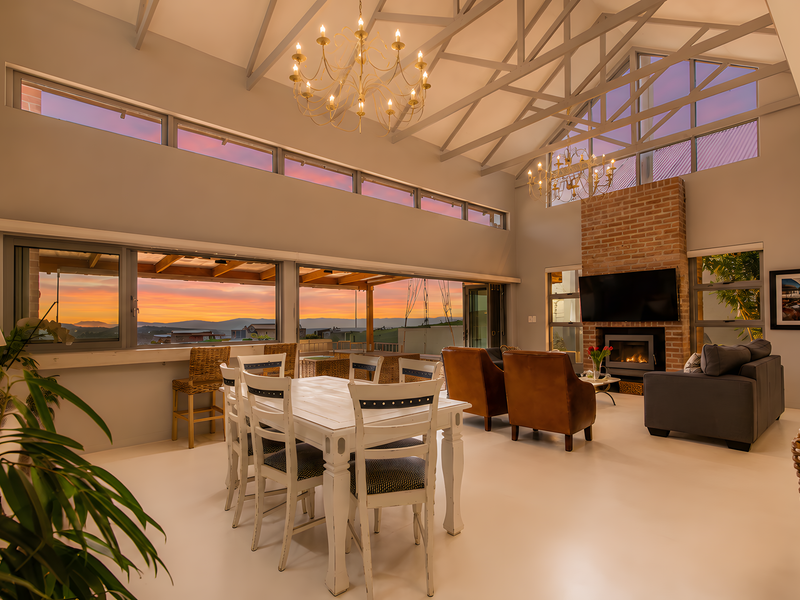 Spacious open-plan living and dining area featuring vaulted ceilings with exposed trusses, a brick fireplace, and large stacking doors opening to scenic views.