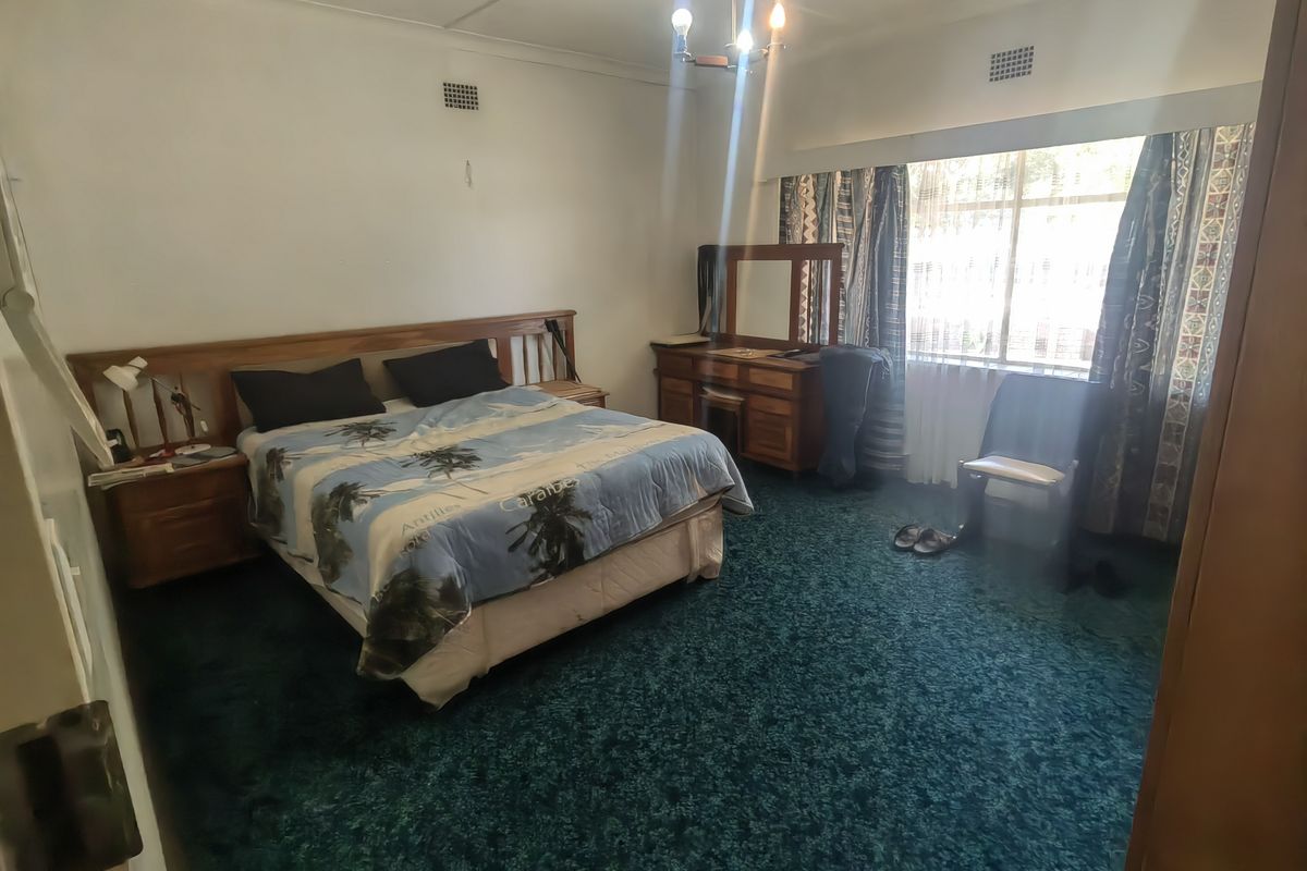 A spacious bedroom featuring dark green carpeting, a large window for natural light, and built-in wooden headboard and bedside cabinetry.