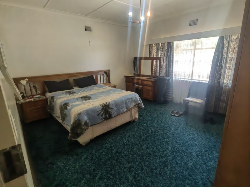 A spacious bedroom featuring dark green carpeting, a large window for natural light, and built-in wooden headboard and bedside cabinetry.