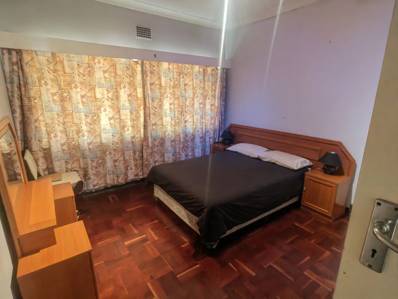 A bedroom featuring parquet flooring, a built-in headboard with integrated bedside tables, and large window coverings.