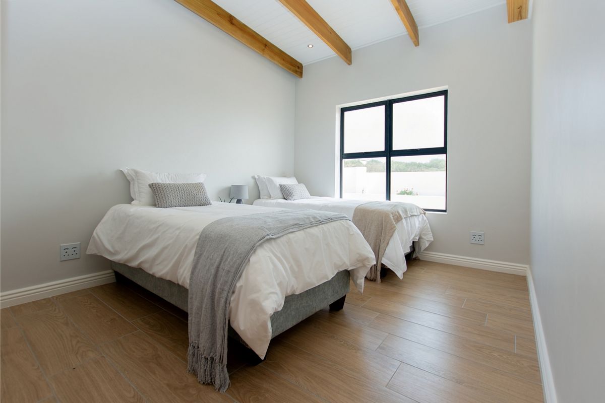 A bright, modern bedroom featuring exposed timber ceiling beams, light wood-look tiled flooring, and a large black-framed window providing natural light.