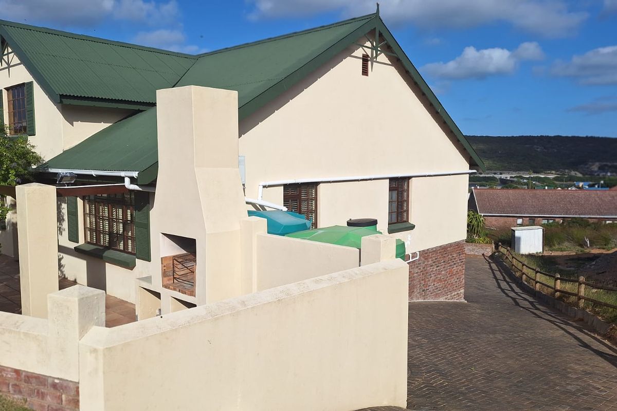 Rear exterior view of the house featuring a built-in braai, paved driveway, water storage tanks, and a pitched corrugated metal roof.
