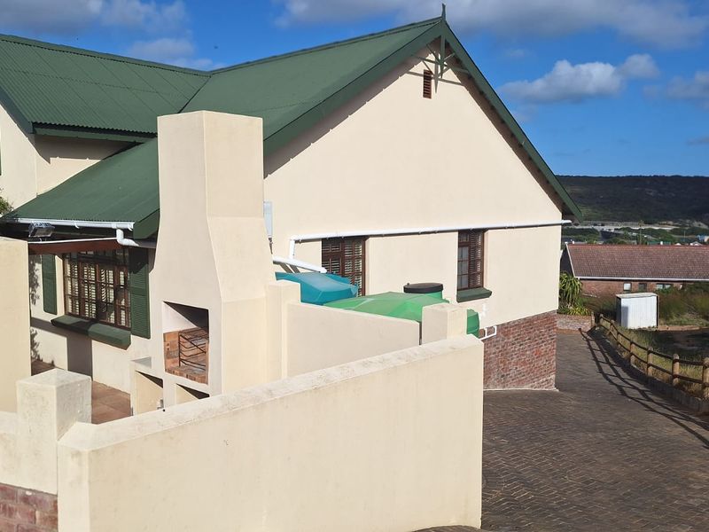 Rear exterior view of the house featuring a built-in braai, paved driveway, water storage tanks, and a pitched corrugated metal roof.