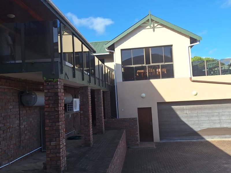 Rear exterior view of the property featuring a paved driveway, integrated garage, and an elevated balcony with glass balustrades supported by brick pillars.