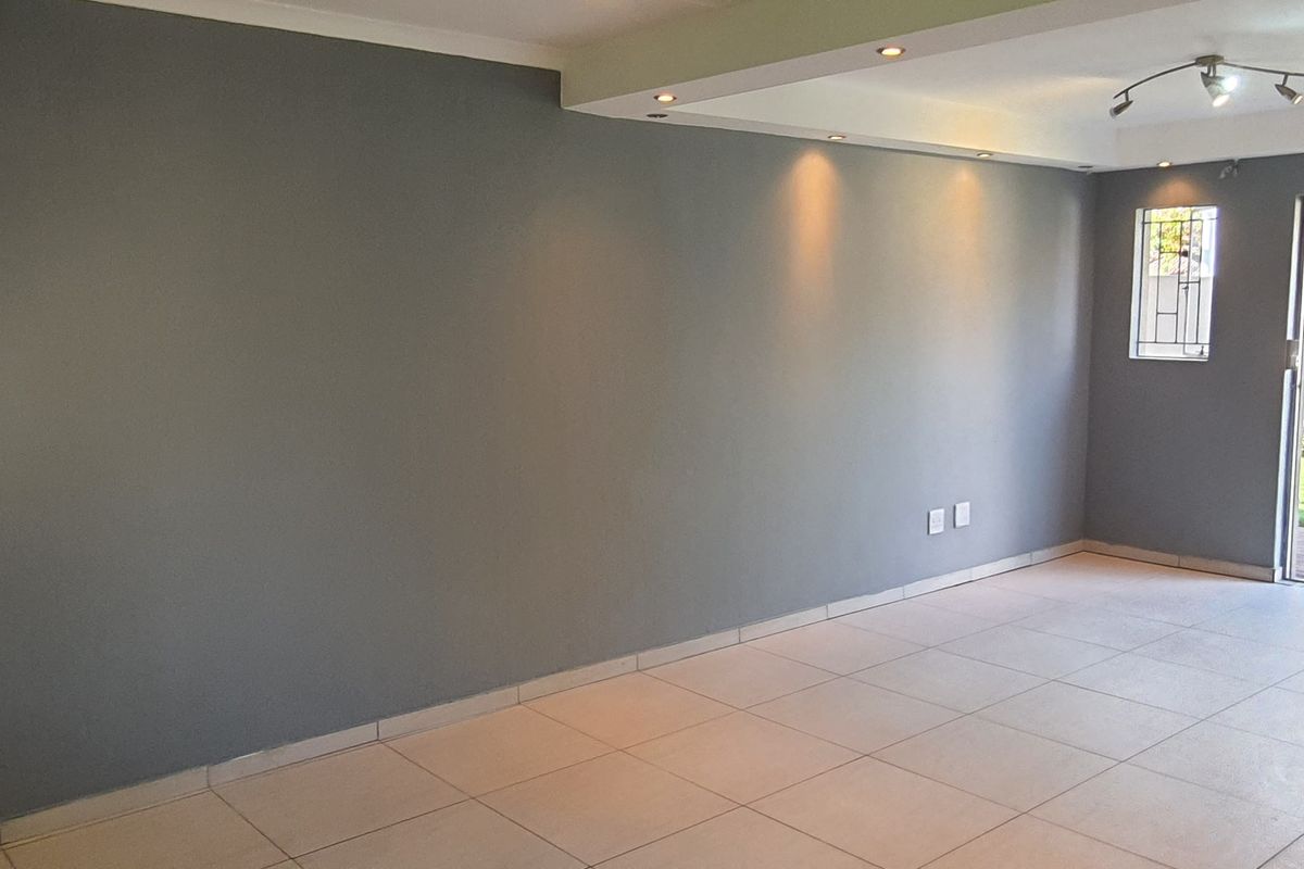 A bright, empty living area featuring neutral grey walls, ceramic floor tiles, recessed lighting, and direct access to an outdoor garden space.
