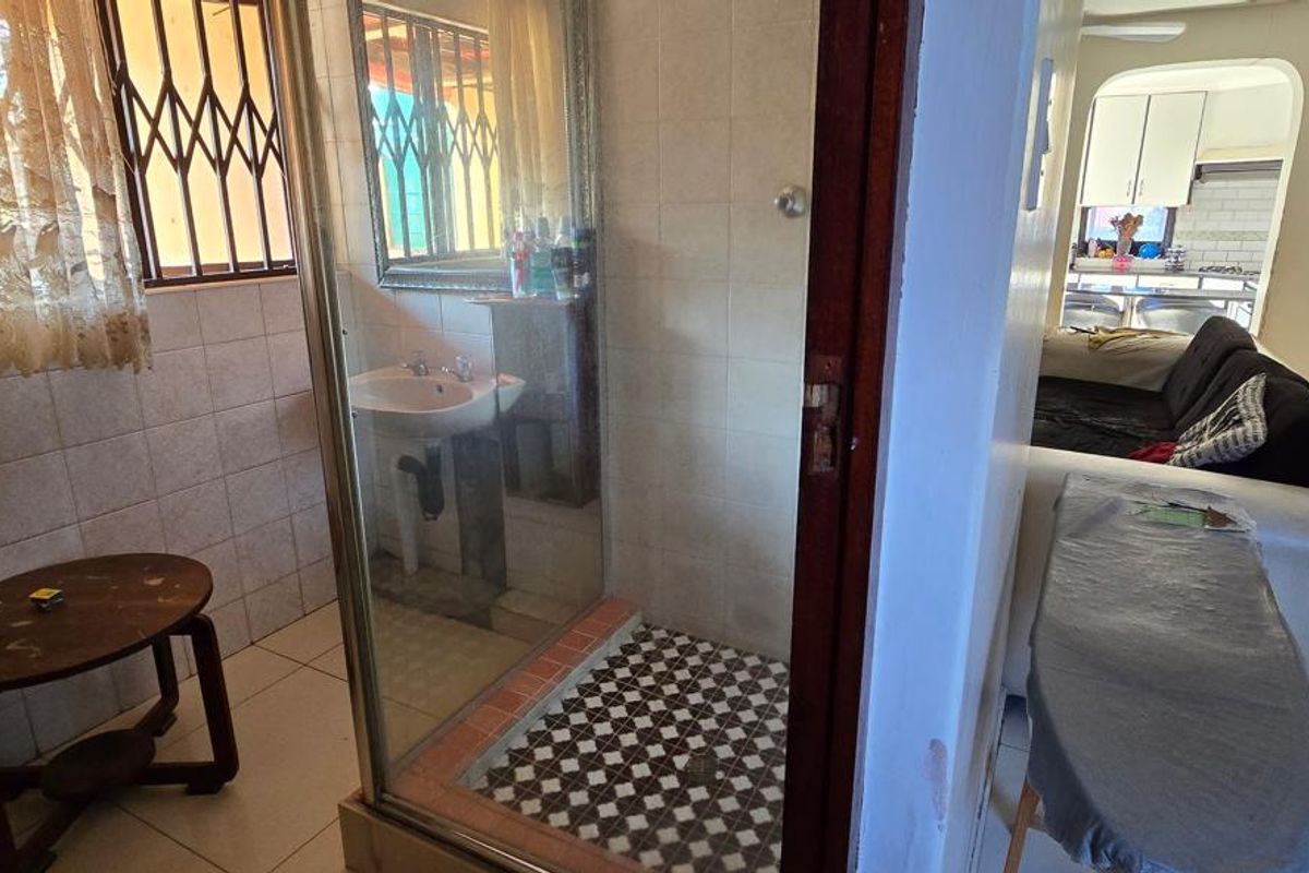 A bathroom featuring a glass-enclosed shower and pedestal basin, with tiled flooring and walls, adjacent to a living area.