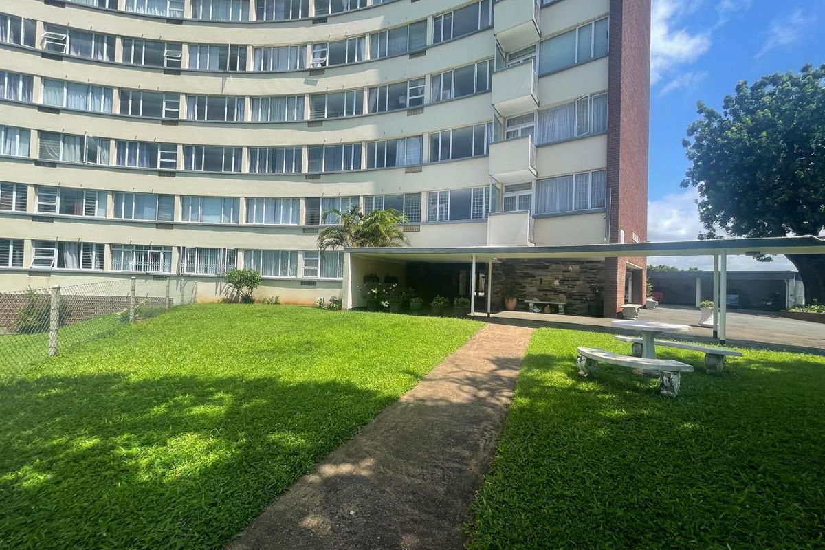 Exterior view of a multi-story apartment building featuring a landscaped lawn, paved pathway, and a covered entrance area with stone detailing.