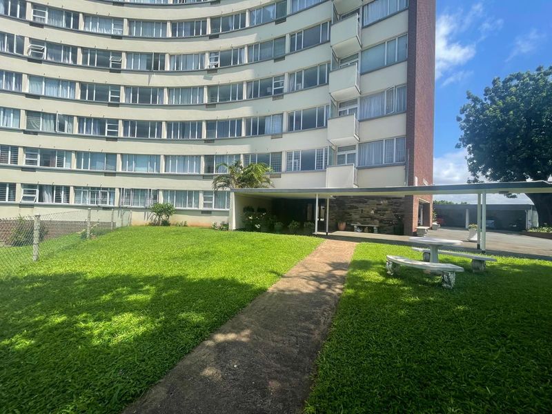 Exterior view of a multi-story apartment building featuring a landscaped lawn, paved pathway, and a covered entrance area with stone detailing.
