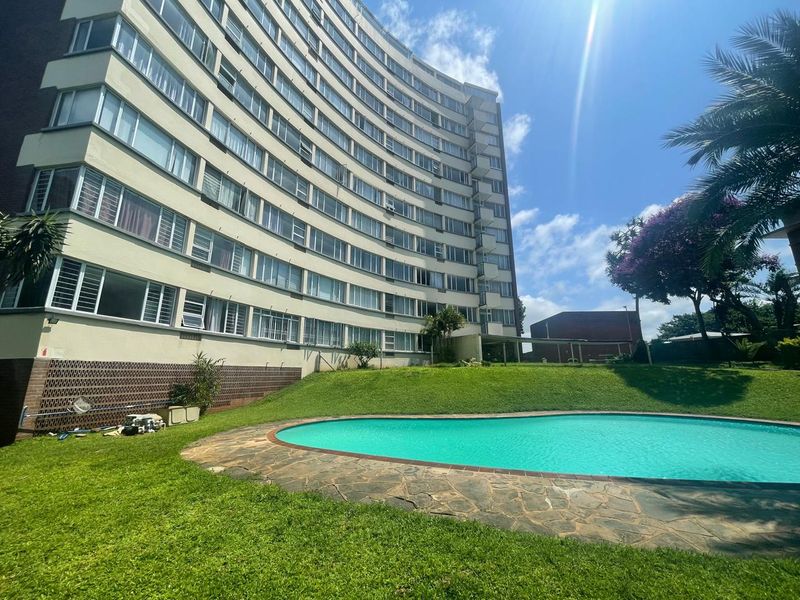 A view of the apartment complex exterior featuring a swimming pool, stone paving, and well-maintained lawn area.