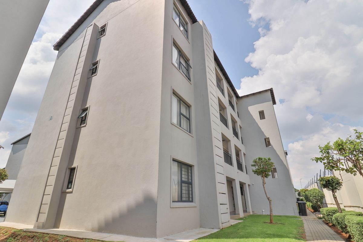 A view of the rear elevation of a multi-story apartment building featuring grey painted exterior walls, aluminium windows, and a paved garden walkway.