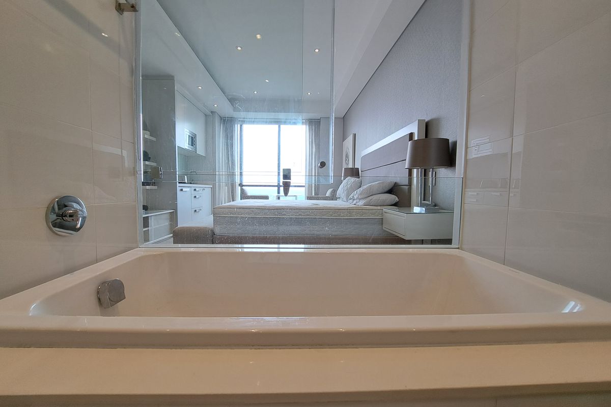 A modern bathroom featuring a built-in bathtub with a glass partition, offering a view into the adjacent bedroom area.