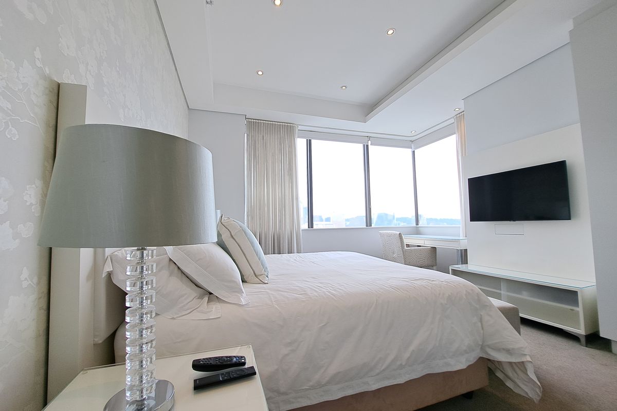 A modern, well-lit bedroom featuring a dropped ceiling with recessed lighting, large windows providing natural light, and a wall-mounted television.