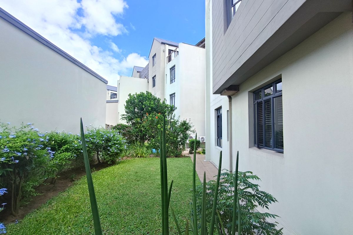 A well-maintained rear garden area featuring a manicured lawn, established shrubs, and a paved walkway between apartment building blocks.