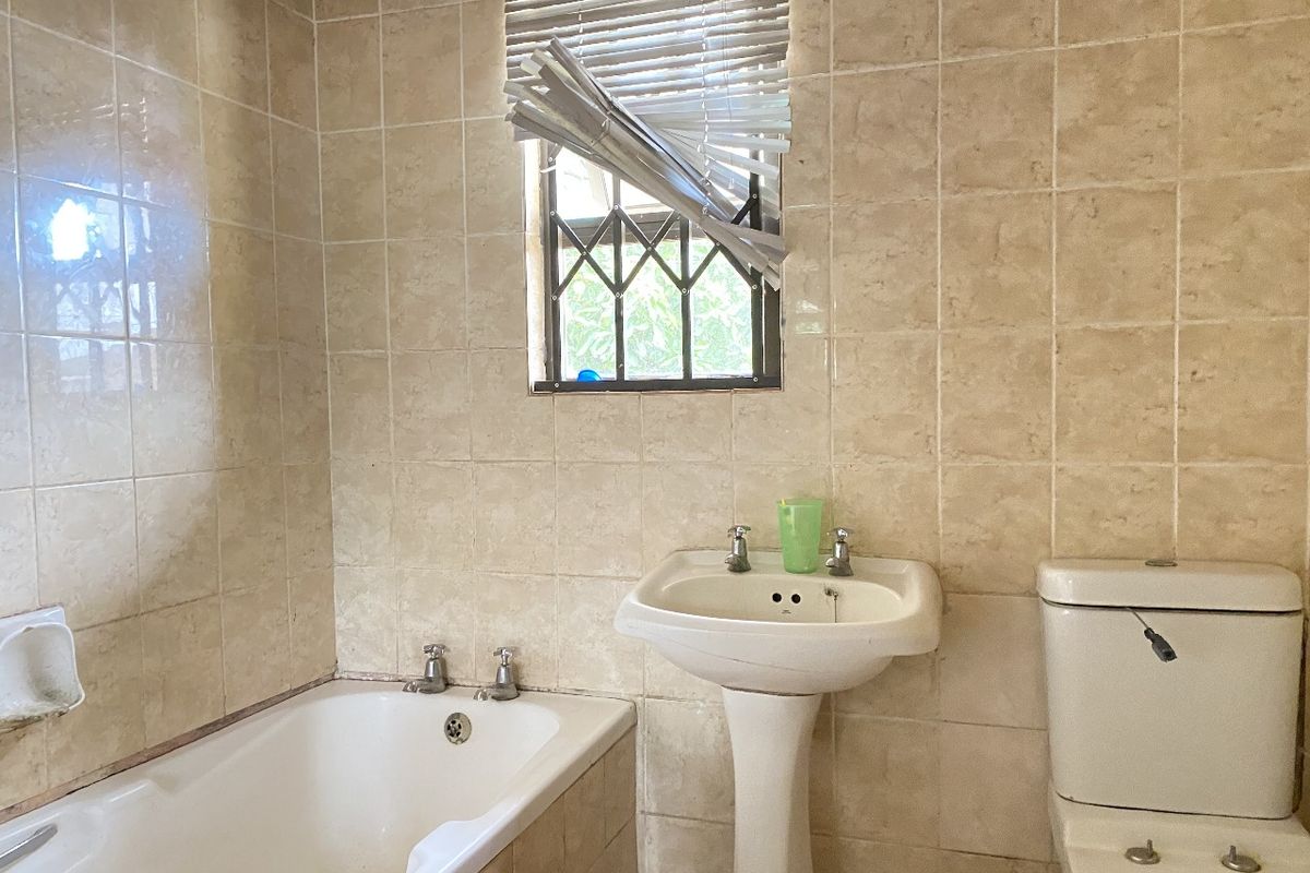 A dated bathroom featuring a standard bathtub, pedestal basin, and toilet, finished with neutral wall tiles and a security-grilled window.