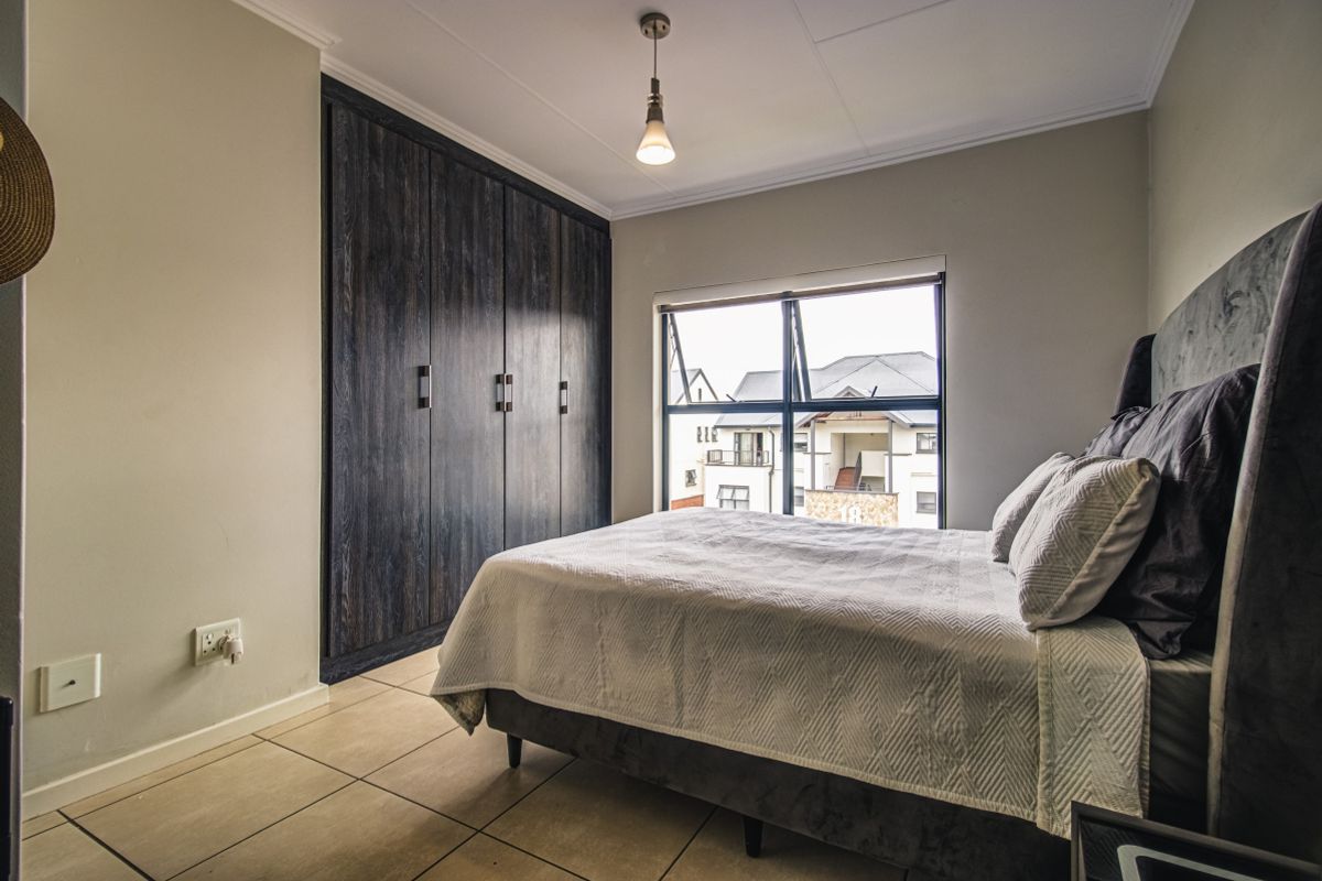 A well-maintained bedroom featuring dark wood built-in cupboards, tiled flooring, and a large window providing natural light and a view of the complex.