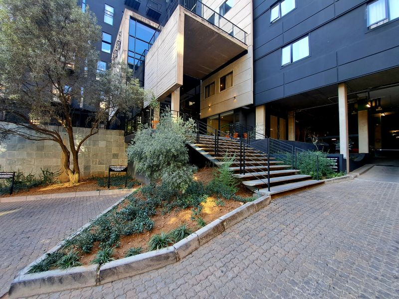 Modern apartment building entrance featuring a paved driveway, concrete staircase with metal railings, and landscaped garden area.