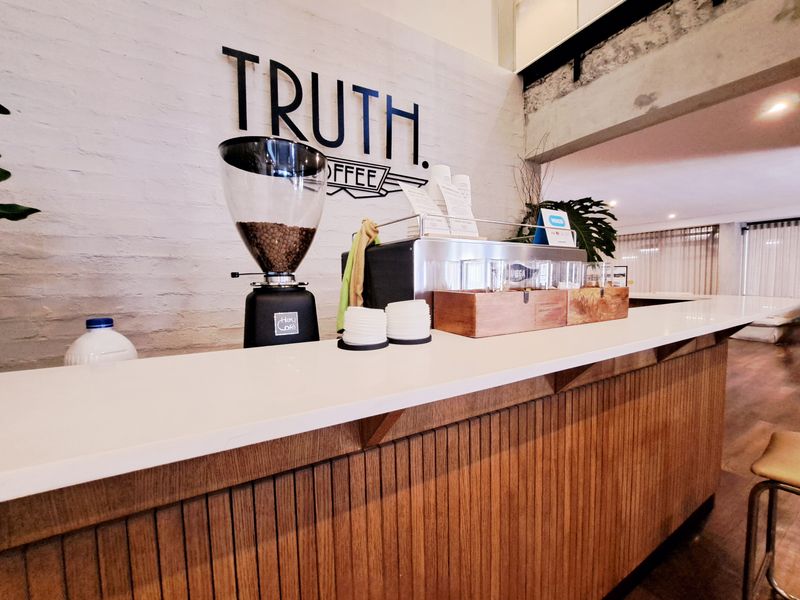 A modern coffee bar area featuring a sleek white countertop, timber slat cabinetry, and an exposed brick wall with branding.
