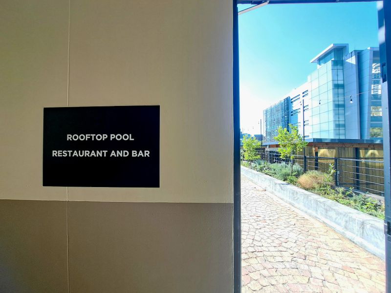 A view from an entrance leading to a rooftop pool, restaurant, and bar area featuring a cobblestone path and modern building surroundings.