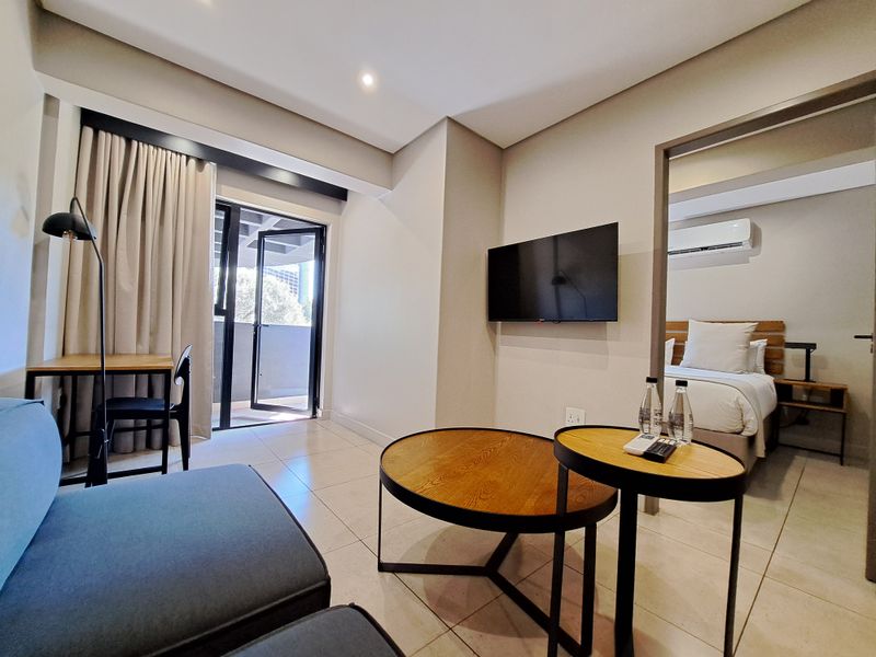 Modern open-plan living area featuring tiled floors, recessed lighting, and a wall-mounted television, with direct access to a private balcony.