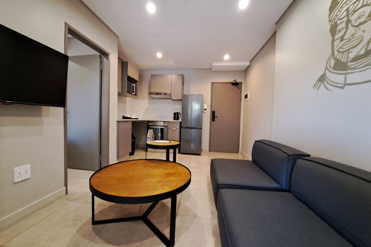 Modern open-plan living area featuring a compact kitchen with built-in cabinetry, oven, and extractor fan, complemented by neutral tiled flooring and recessed lighting.
