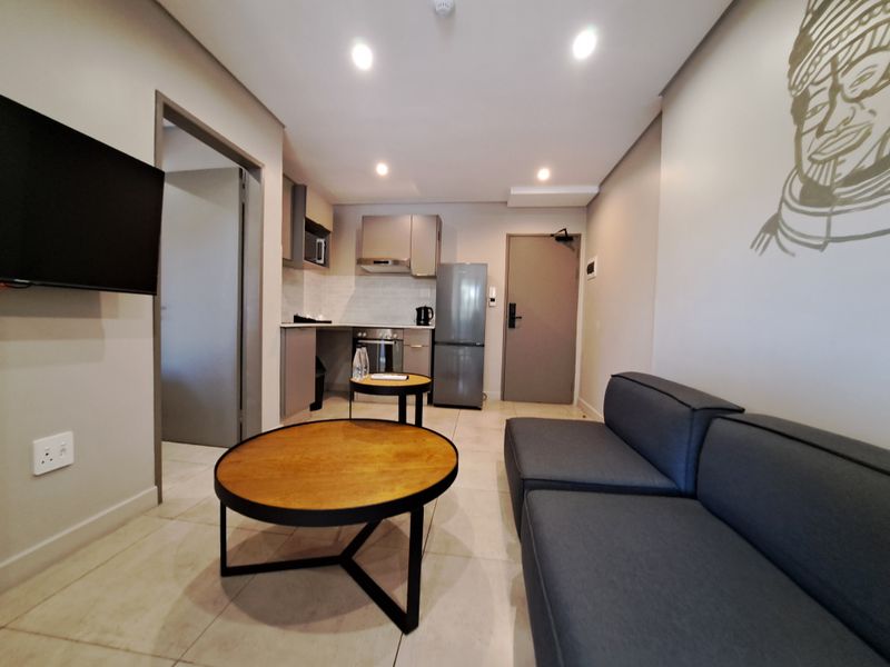 Modern open-plan living area featuring a compact kitchen with built-in cabinetry, oven, and extractor fan, complemented by neutral tiled flooring and recessed lighting.